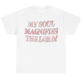 A white short-sleeve crewneck t-shirt featuring the text 'MY SOUL MAGNIFIES THE LORD!' printed on the chest in a textured, distressed pink font.