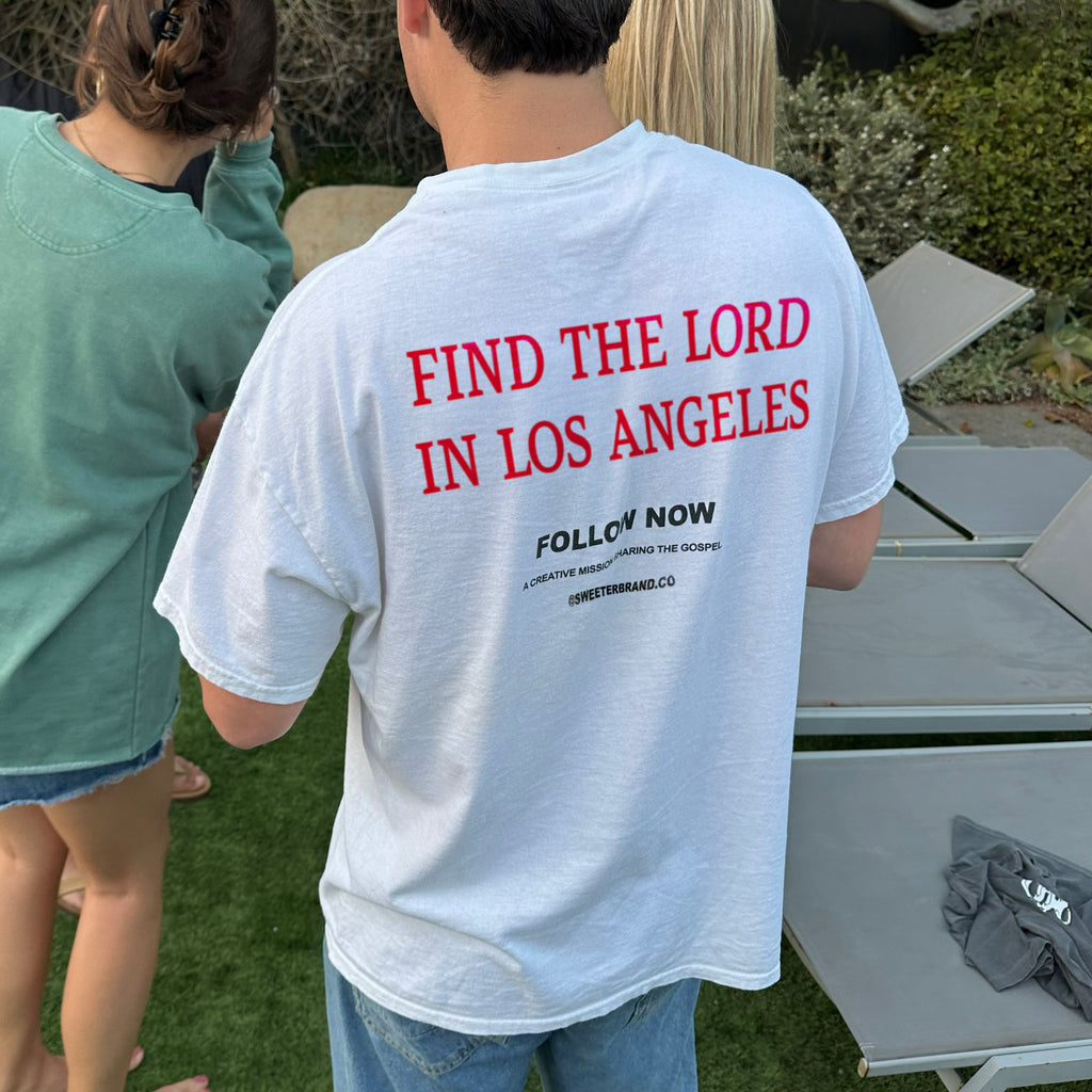 A person standing outdoors on grass wearing a white T-shirt with bold red text reading 'FIND THE LORD IN LOS ANGELES' and smaller black text below, photographed from the back. Other people and lounge chairs are visible in the background.