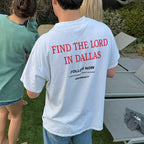 A person standing outdoors on grass wearing a white T-shirt with bold red text reading 'FIND THE LORD IN DALLAS' and smaller black text below, photographed from the back. Other people and lounge chairs are visible in the background.