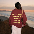 A person standing outdoors at sunset facing away from the camera, wearing an oversized burgundy sweatshirt with large yellow text on the back that reads 'TELL THE WORLD THAT JESUS SAVES', paired with white pants and a shoulder bag.