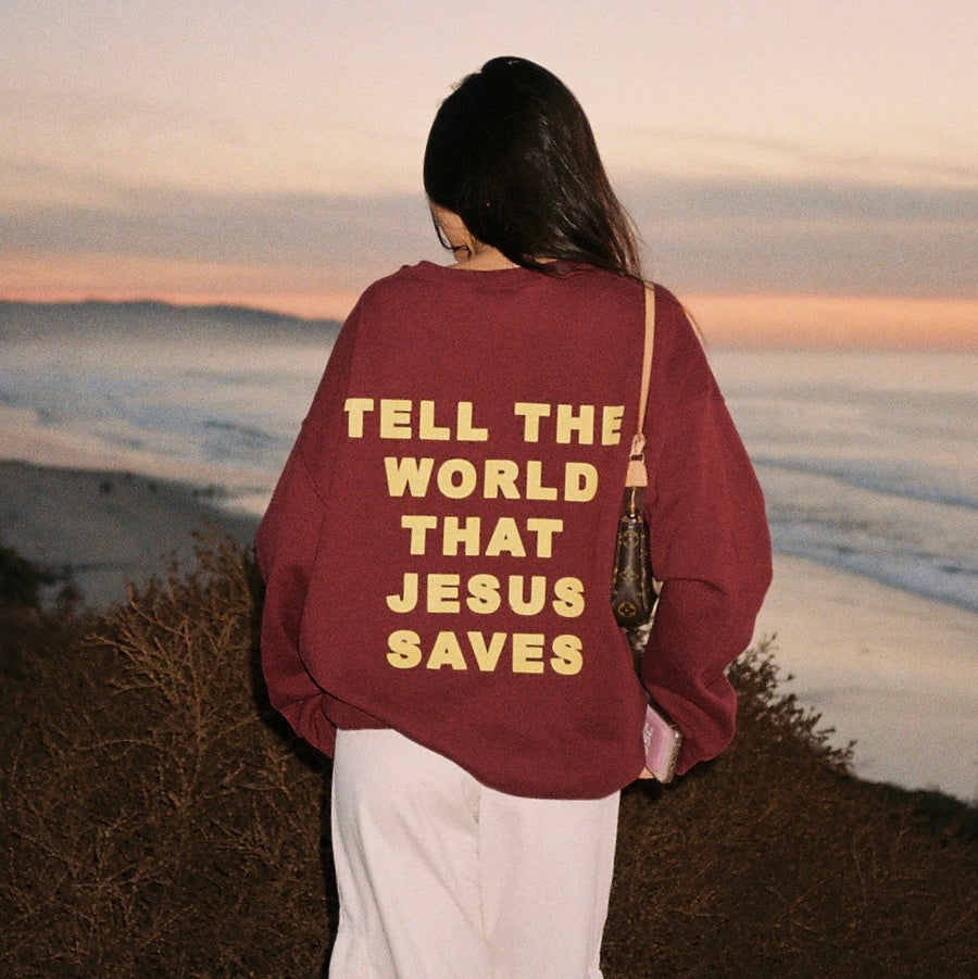 A person standing outdoors at sunset facing away from the camera, wearing an oversized burgundy sweatshirt with large yellow text on the back that reads 'TELL THE WORLD THAT JESUS SAVES', paired with white pants and a shoulder bag.
