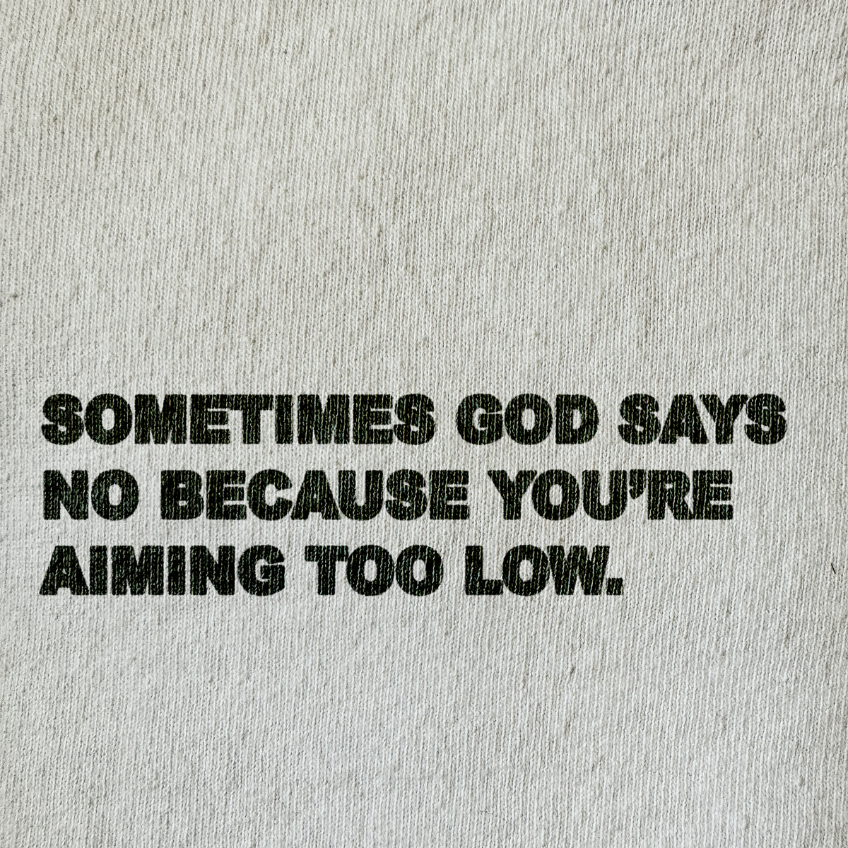 Black distressed text reading 'SOMETIMES GOD SAYS NO BECAUSE YOU'RE AIMING TOO LOW.' printed on a light beige textured fabric background.