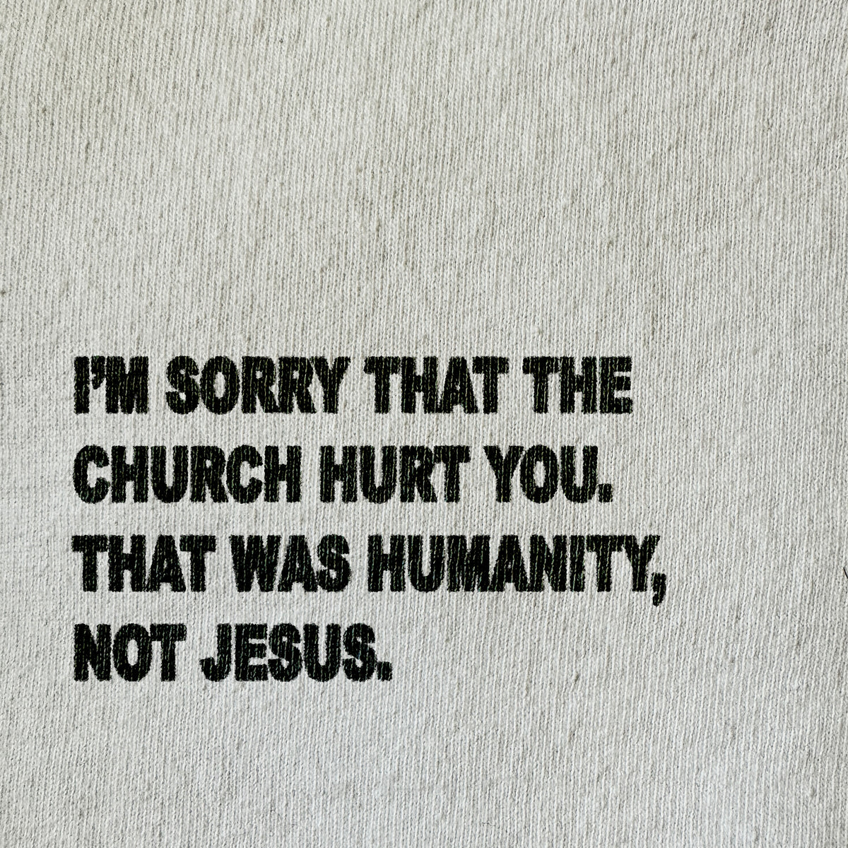 A close-up of a light beige textured fabric with bold black printed text that reads, 'I'M SORRY THAT THE CHURCH HURT YOU. THAT WAS HUMANITY, NOT JESUS.