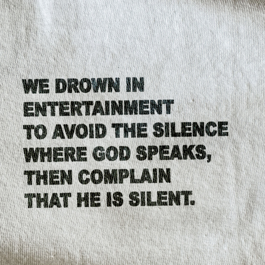 Close-up of a light gray or off-white fabric surface with bold black printed text reading, 'WE DROWN IN ENTERTAINMENT TO AVOID THE SILENCE WHERE GOD SPEAKS, THEN COMPLAIN THAT HE IS SILENT.'.