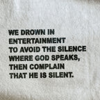 Close-up of a light gray or off-white fabric surface with bold black printed text reading, 'WE DROWN IN ENTERTAINMENT TO AVOID THE SILENCE WHERE GOD SPEAKS, THEN COMPLAIN THAT HE IS SILENT.'.