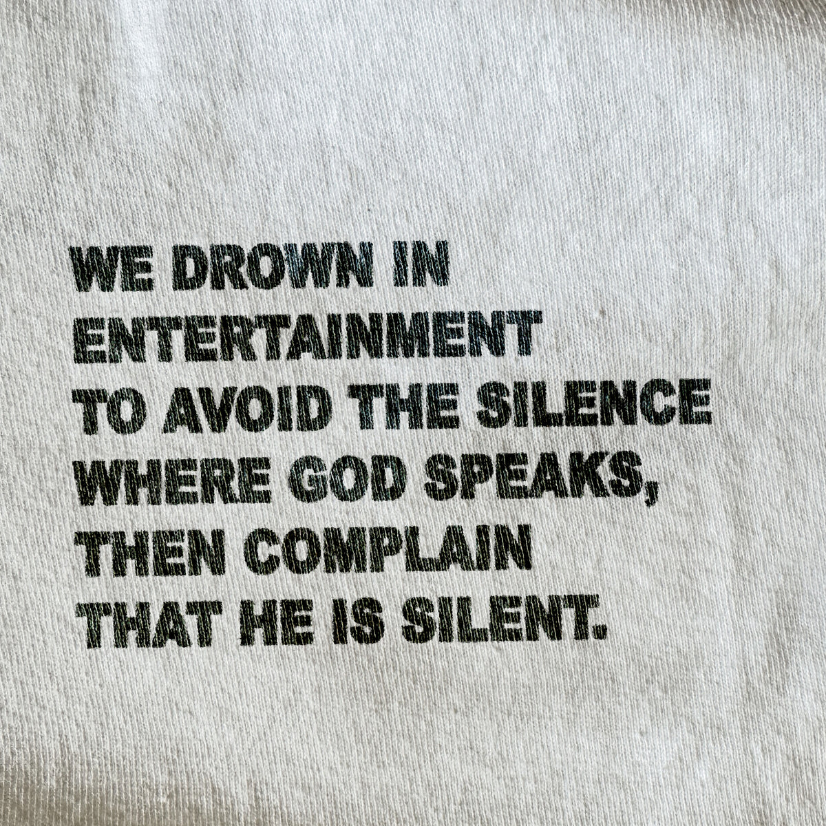Close-up of a light gray or off-white fabric surface with bold black printed text reading, 'WE DROWN IN ENTERTAINMENT TO AVOID THE SILENCE WHERE GOD SPEAKS, THEN COMPLAIN THAT HE IS SILENT.'.