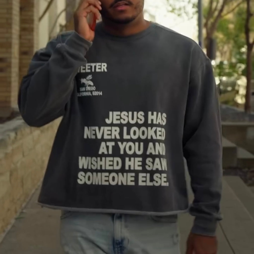 A man wearing a dark gray crewneck sweatshirt with bold white text that reads 'JESUS HAS NEVER LOOKED AT YOU AND WISHED HE SAW SOMEONE ELSE.' He is standing outdoors on a sidewalk, holding a phone to his ear.