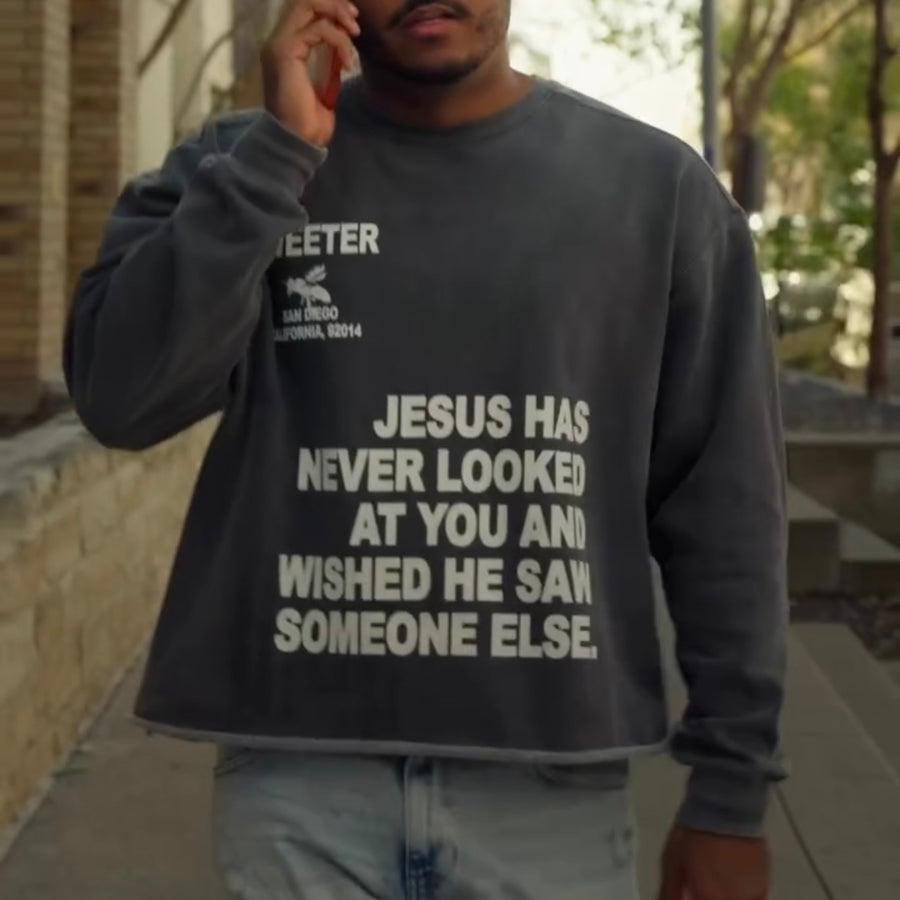A man wearing a dark gray crewneck sweatshirt with bold white text that reads 'JESUS HAS NEVER LOOKED AT YOU AND WISHED HE SAW SOMEONE ELSE.' He is standing outdoors on a sidewalk, holding a phone to his ear.