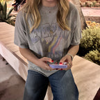 A woman wearing a gray graphic t-shirt with a rainbow design and the text 'NEVER HIDE', paired with blue jeans, sitting on a wooden bench outdoors.