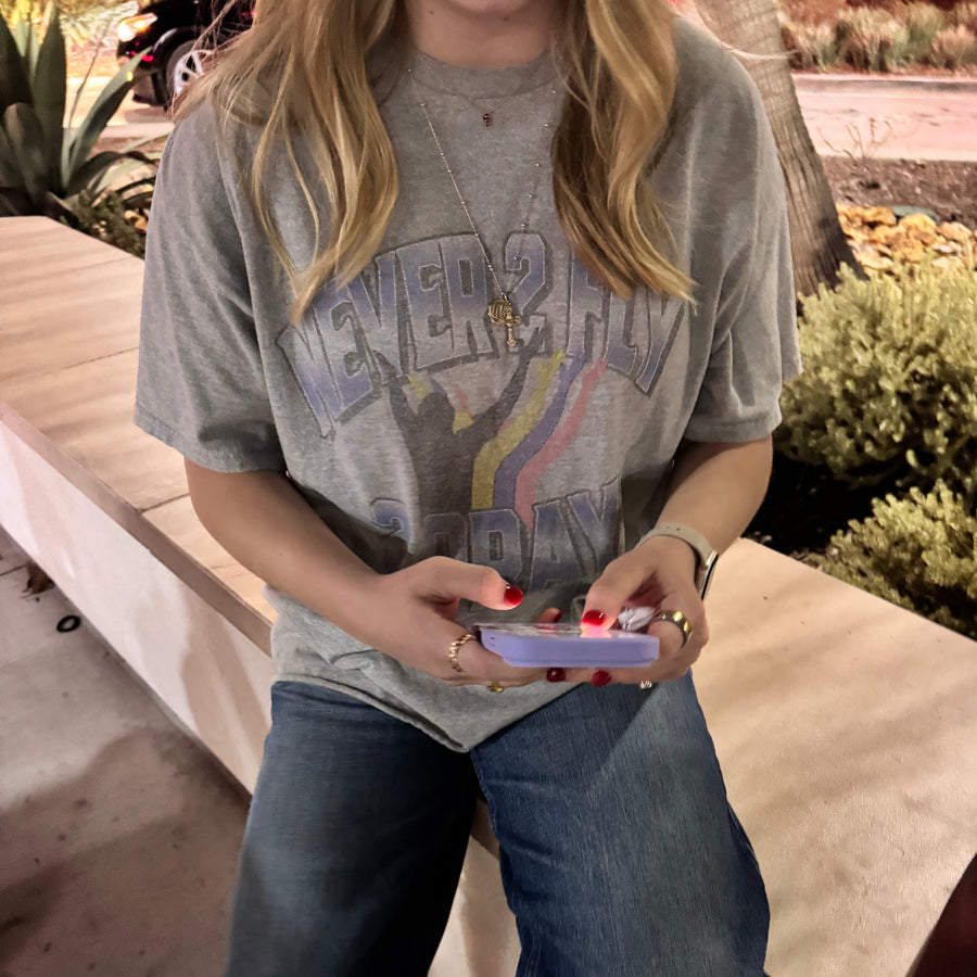 A woman wearing a gray graphic t-shirt with a rainbow design and the text 'NEVER HIDE', paired with blue jeans, sitting on a wooden bench outdoors.