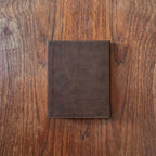 A dark brown leather notebook cover or tablet sleeve, shown closed from the front on a rustic wooden surface.