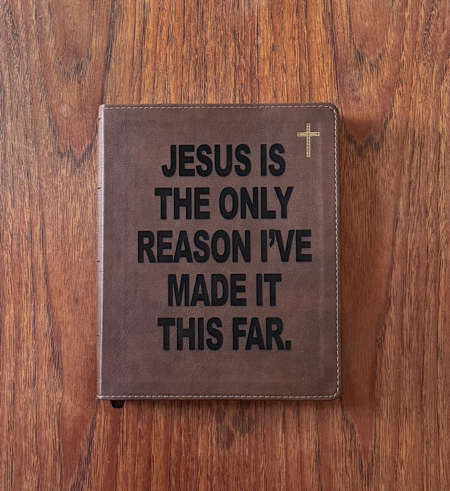 A brown leather Bible cover or journal lying flat on a wooden table, featuring the text 'JESUS IS THE ONLY REASON I'VE MADE IT THIS FAR.' in black lettering, a small gold cross at the top right, and visible stitching around the edges.