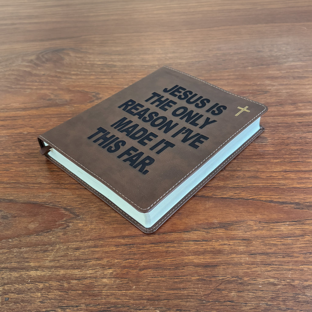 A brown leather-bound book with the text 'JESUS IS THE ONLY REASON I'VE MADE IT THIS FAR.' in dark lettering, a small gold cross, and light blue pages visible on the side, placed on a wooden tabletop.
