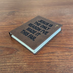 A brown leather-bound book with the text 'JESUS IS THE ONLY REASON I'VE MADE IT THIS FAR.' in dark lettering, a small gold cross, and light blue pages visible on the side, placed on a wooden tabletop.