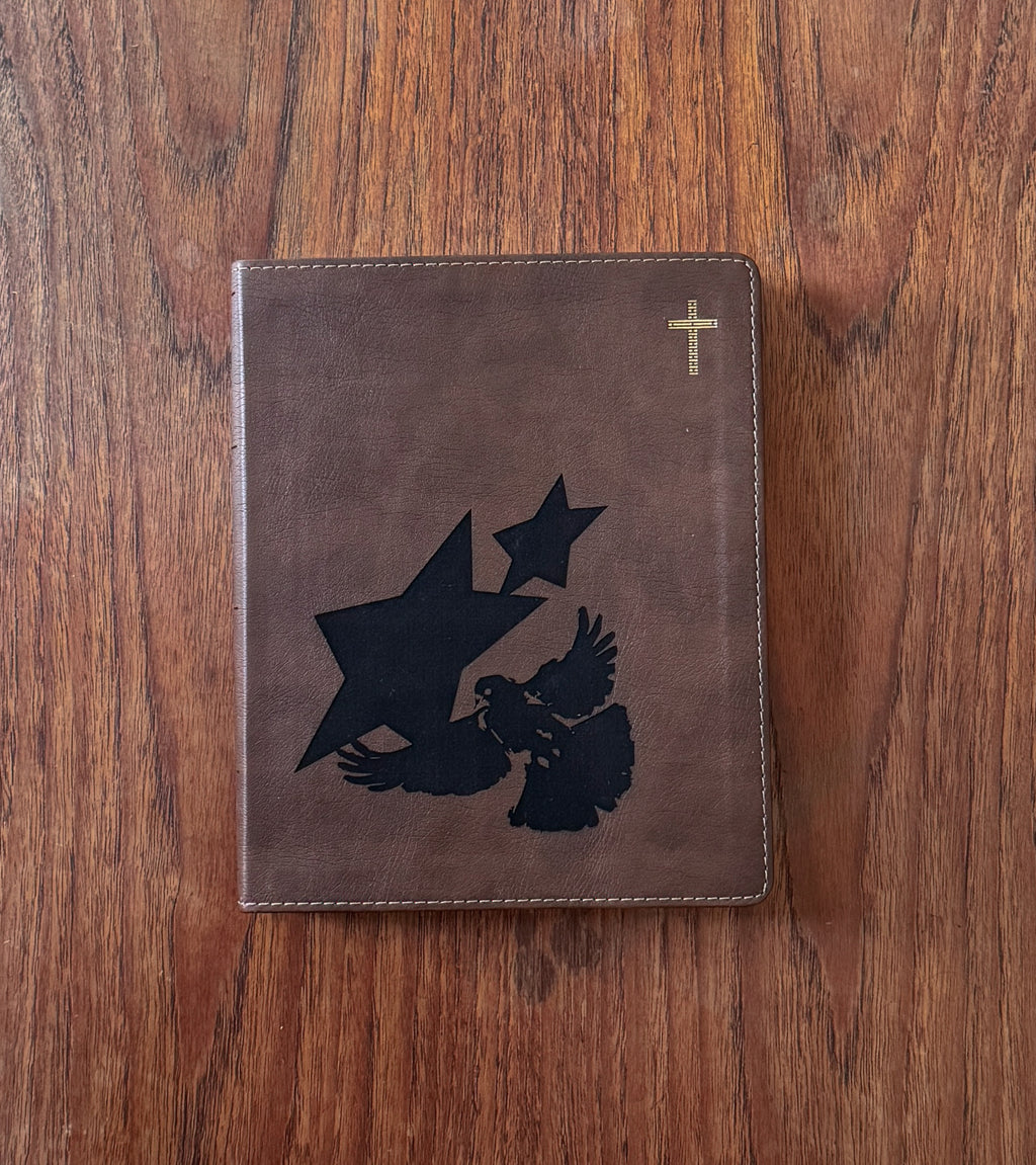 A brown leather Bible or prayer journal with a black eagle and star design on the front and a small gold cross in the top right corner, displayed on a wooden surface.