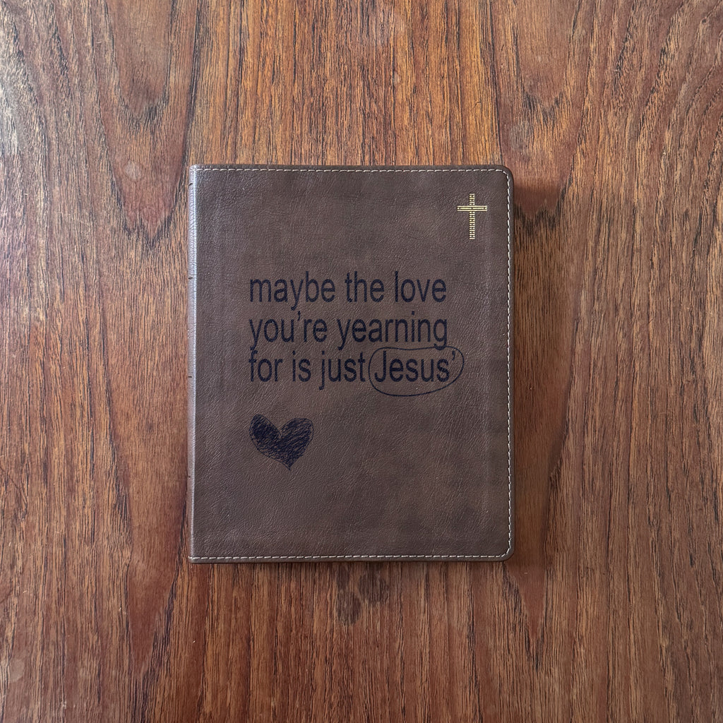 A brown leather Bible cover or journal lying flat on a wooden tabletop, with embossed text reading 'maybe the love you're yearning for is just Jesus,' a small cross near the top right, and a heart near the bottom left.