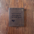 A brown leather Bible cover or journal lying flat on a wooden tabletop, with embossed text reading 'maybe the love you're yearning for is just Jesus,' a small cross near the top right, and a heart near the bottom left.