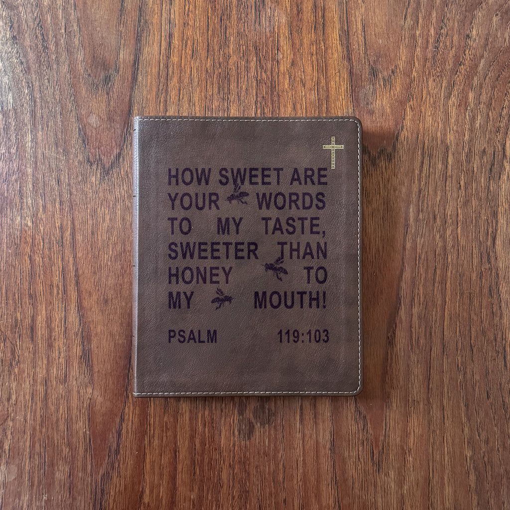 A brown leather Bible cover featuring an engraved quote from Psalm 119:103 and a small gold cross, displayed flat on a wooden surface.