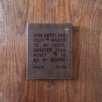 A brown leather Bible cover featuring an engraved quote from Psalm 119:103 and a small gold cross, displayed flat on a wooden surface.