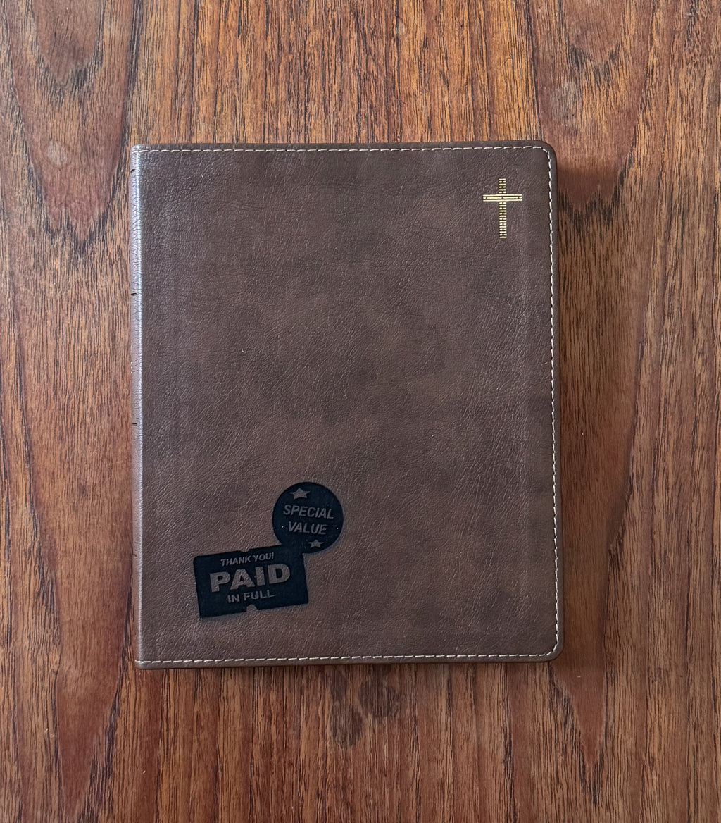 A brown faux-leather Bible cover or journal with a gold cross in the top right corner, placed on a wooden table. A black 'Thank You Paid in Full' card is tucked into the bottom left pocket.