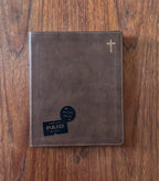 A brown faux-leather Bible cover or journal with a gold cross in the top right corner, placed on a wooden table. A black 'Thank You Paid in Full' card is tucked into the bottom left pocket.