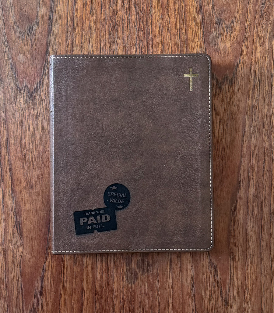 A brown faux-leather Bible cover or journal with a gold cross in the top right corner, placed on a wooden table. A black 'Thank You Paid in Full' card is tucked into the bottom left pocket.