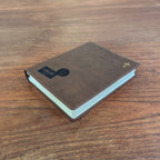 A brown leather-bound book with a gold cross on the cover, placed on a wooden table. A black and blue 'PAID' card is attached to the front cover.
