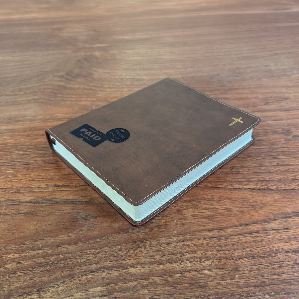 A brown leather-bound book with a gold cross on the cover, placed on a wooden table. A black and blue 'PAID' card is attached to the front cover.