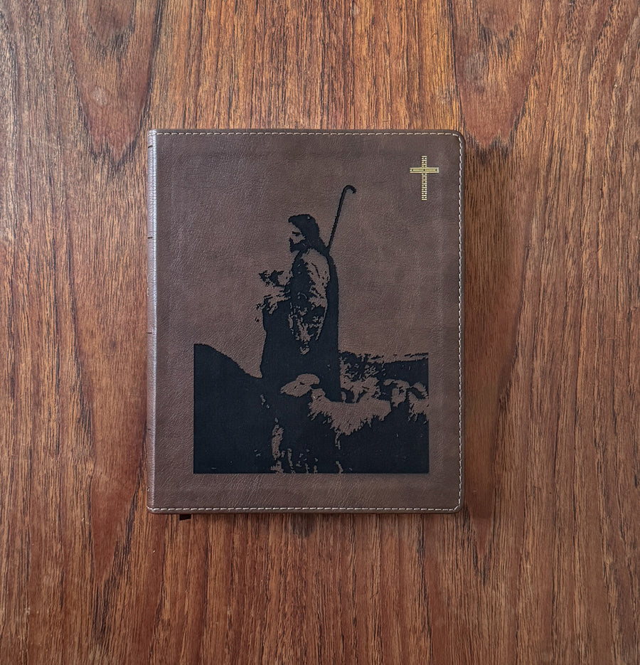 A brown faux-leather Bible or prayer journal with a black graphic of a shepherd and sheep on the front cover, featuring a small gold cross in the top right corner, displayed on a wooden table.