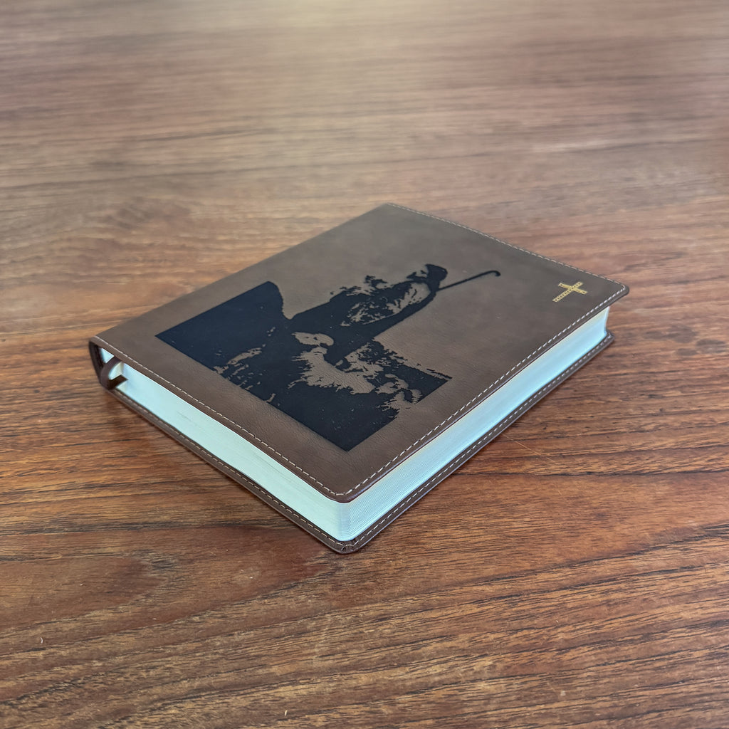 A brown leather-bound book with a black silhouette design on the cover and a small gold cross on the front, photographed on a wooden tabletop.