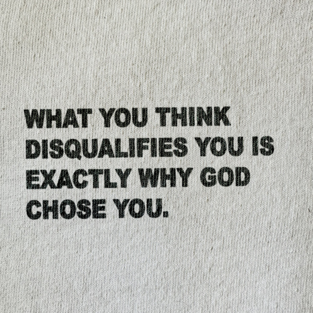 A close-up of a light beige or off-white textured fabric, likely a t-shirt or sweatshirt, featuring a bold black printed typographic quote that reads: 'WHAT YOU THINK DISQUALIFIES YOU IS EXACTLY WHY GOD CHOSE YOU.'.