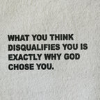 A close-up of a light beige or off-white textured fabric, likely a t-shirt or sweatshirt, featuring a bold black printed typographic quote that reads: 'WHAT YOU THINK DISQUALIFIES YOU IS EXACTLY WHY GOD CHOSE YOU.'.