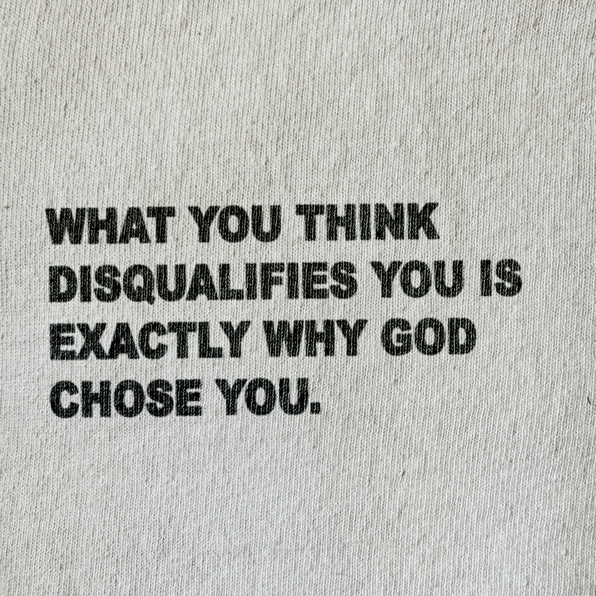 A close-up of a light beige or off-white textured fabric, likely a t-shirt or sweatshirt, featuring a bold black printed typographic quote that reads: 'WHAT YOU THINK DISQUALIFIES YOU IS EXACTLY WHY GOD CHOSE YOU.'.