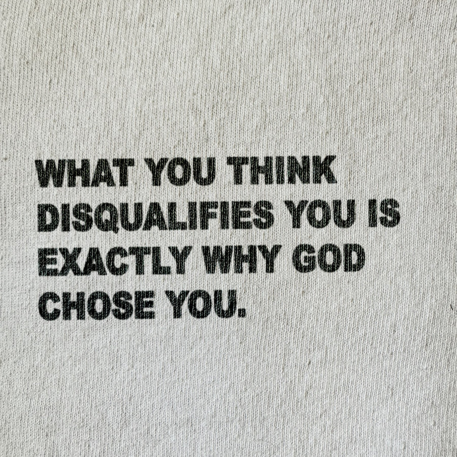 A close-up of a light beige or off-white textured fabric, likely a t-shirt or sweatshirt, featuring a bold black printed typographic quote that reads: 'WHAT YOU THINK DISQUALIFIES YOU IS EXACTLY WHY GOD CHOSE YOU.'.