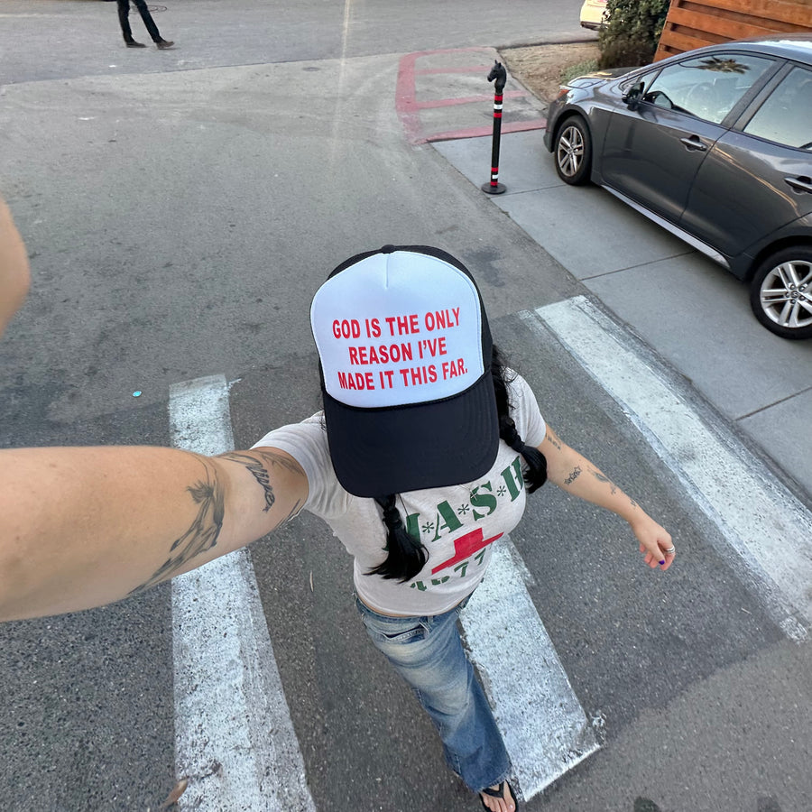 A person taking a selfie in a crosswalk wearing a white and navy trucker hat with red text that reads 'GOD IS THE ONLY REASON I'VE MADE IT THIS FAR'.