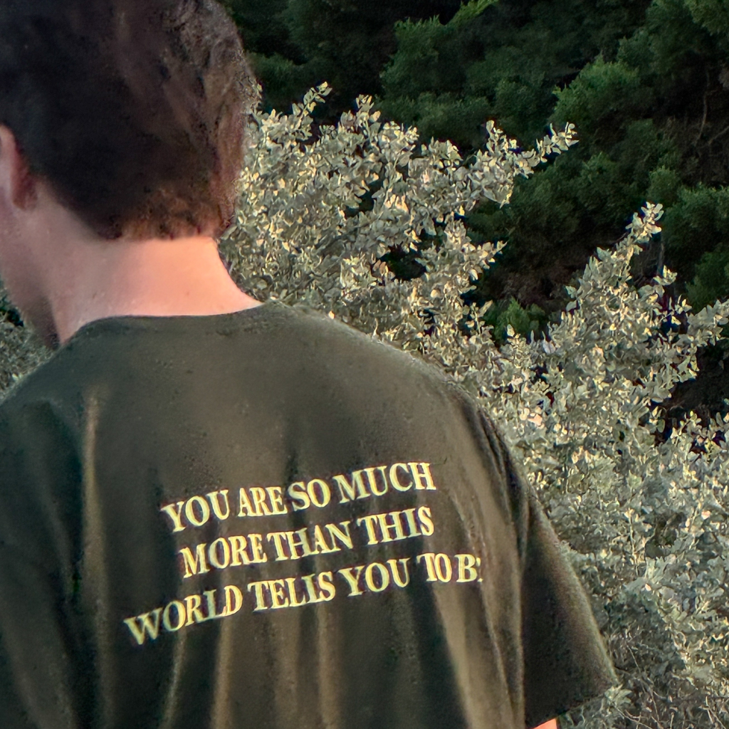 A person viewed from behind wearing an olive green t-shirt with yellow text that reads 'YOU ARE SO MUCH MORE THAN THIS WORLD TELIS YOU TO B.' outdoors in front of green foliage.