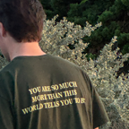 A person viewed from behind wearing an olive green t-shirt with yellow text that reads 'YOU ARE SO MUCH MORE THAN THIS WORLD TELIS YOU TO B.' outdoors in front of green foliage.