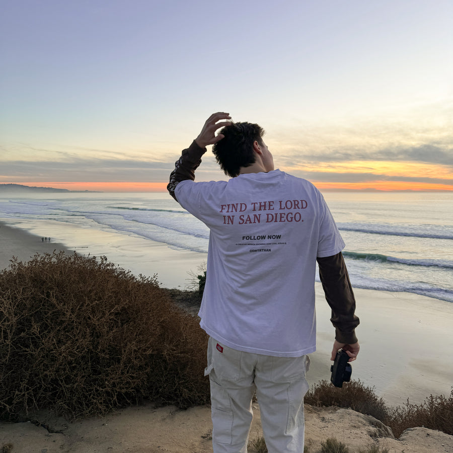 A man stands on a beach cliff at sunset facing the ocean, wearing a white graphic T-shirt with the text 'FIND THE LORD IN SAN DIEGO' and 'FOLLOW NOW' printed on the back, paired with white pants.