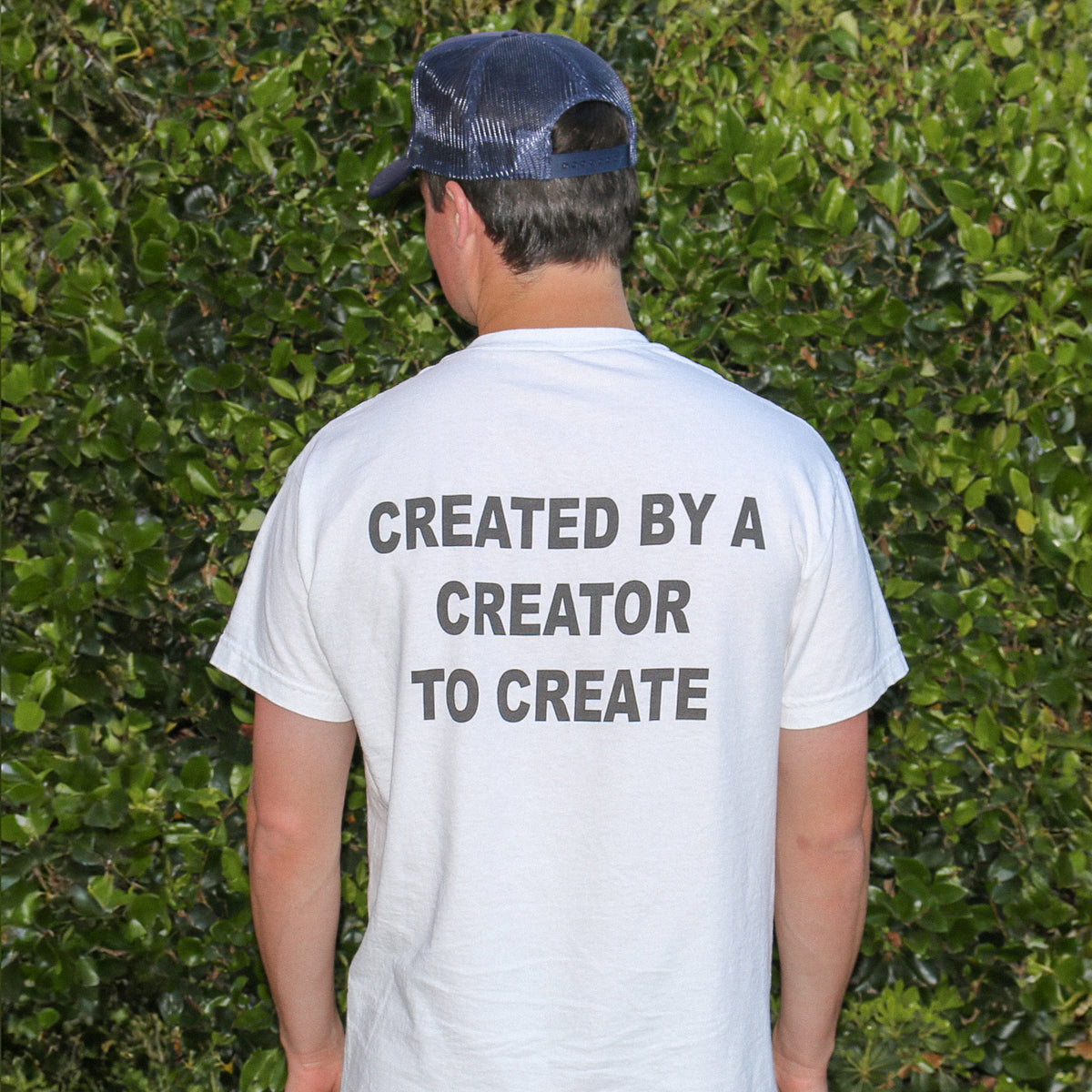 A man viewed from behind wearing a white t-shirt with the text 'CREATED BY A CREATOR TO CREATE' printed in dark gray on the back, paired with a navy blue trucker hat, set against a background of green foliage.