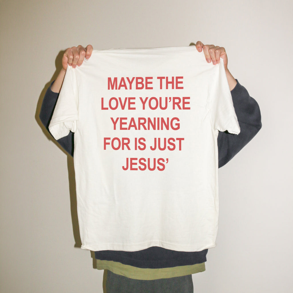 A person holding up a white t-shirt with red text that reads 'MAYBE THE LOVE YOU'RE YEARNING FOR IS JUST JESUS' against a plain beige wall.