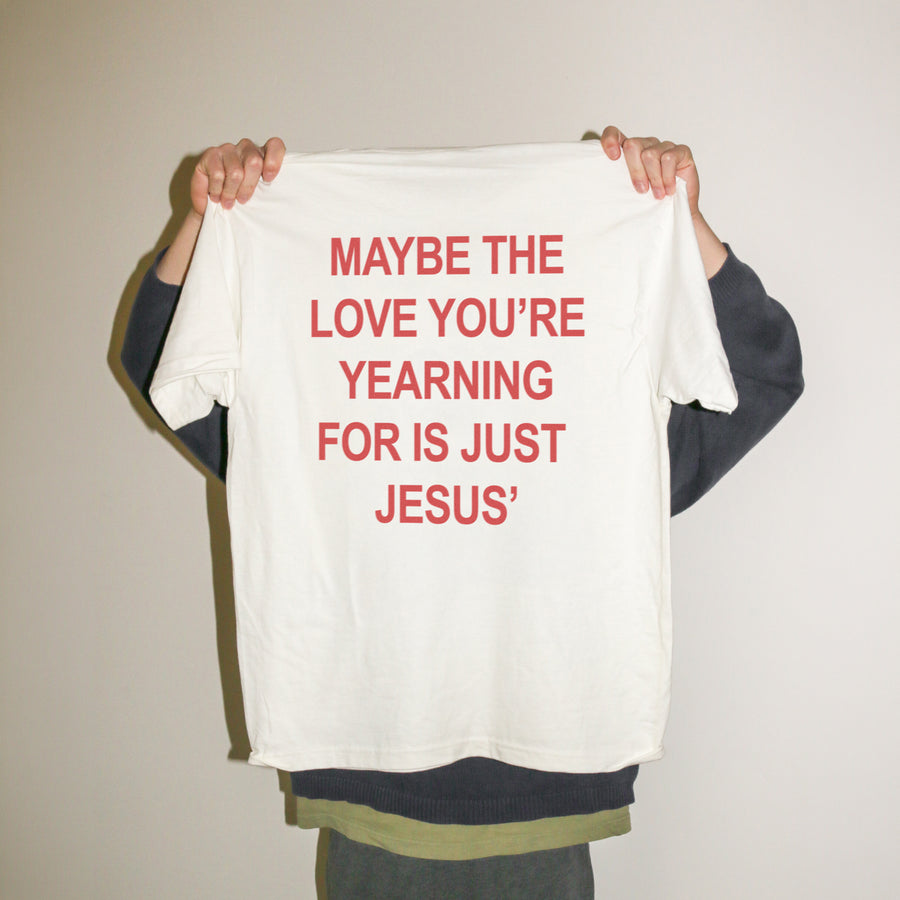 A person holding up a white t-shirt with red text that reads 'MAYBE THE LOVE YOU'RE YEARNING FOR IS JUST JESUS' against a plain beige wall.