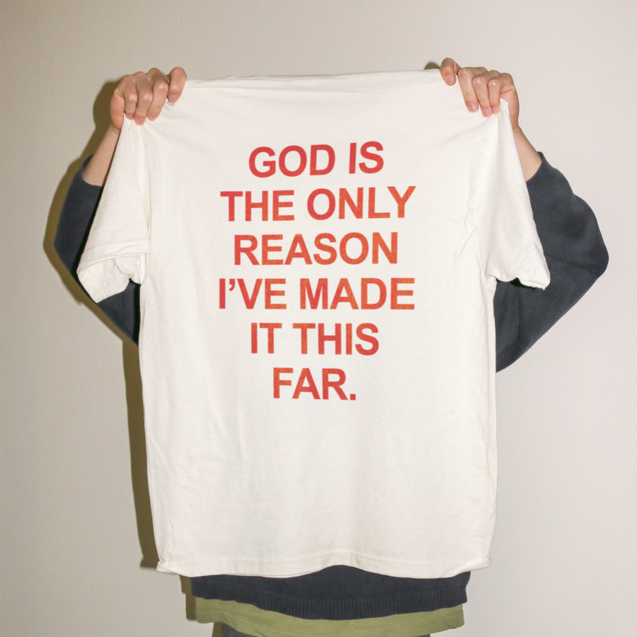 A person holding up a white oversized T-shirt with bold red text that reads, 'GOD IS THE ONLY REASON I’VE MADE IT THIS FAR.' against a plain light beige wall.