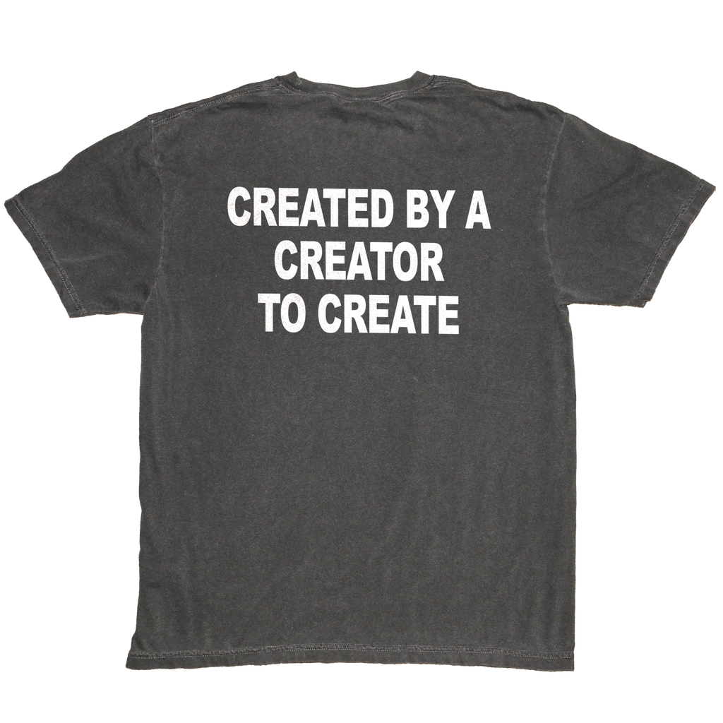 A dark gray washed t-shirt shown from the back, featuring the phrase 'CREATED BY A CREATOR TO CREATE' printed in bold white capital letters across the upper back.