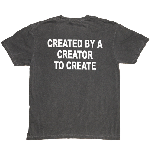 A dark gray washed t-shirt shown from the back, featuring the phrase 'CREATED BY A CREATOR TO CREATE' printed in bold white capital letters across the upper back.