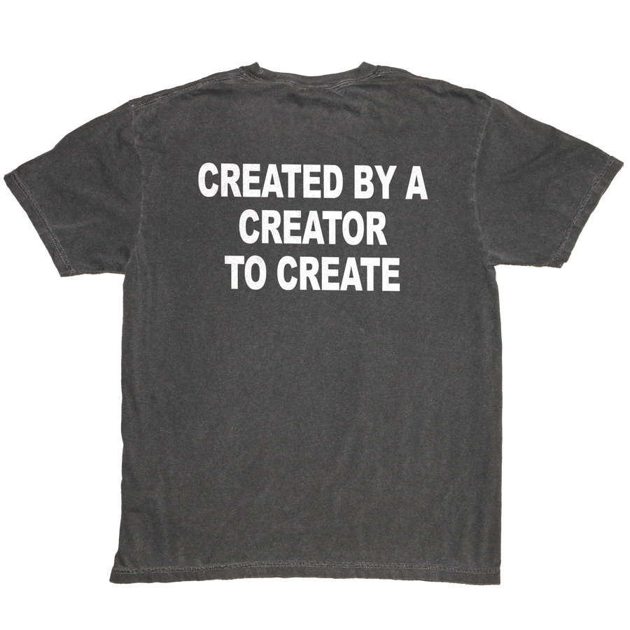 A dark gray washed t-shirt shown from the back, featuring the phrase 'CREATED BY A CREATOR TO CREATE' printed in bold white capital letters across the upper back.