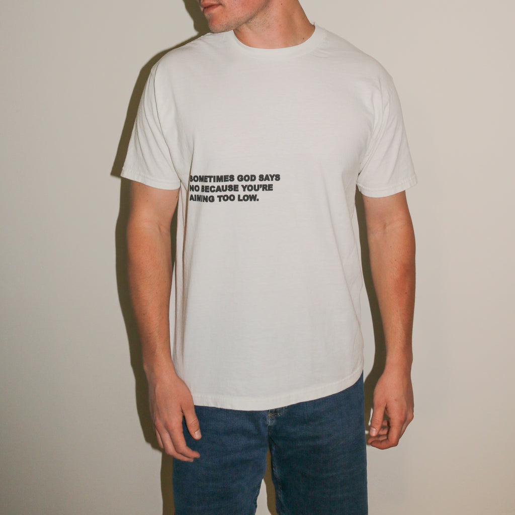 A man wearing a white crewneck t-shirt with black printed text on the chest, paired with blue jeans, standing against a plain beige wall.