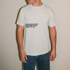 A man wearing a white crewneck t-shirt with black printed text on the chest, paired with blue jeans, standing against a plain beige wall.