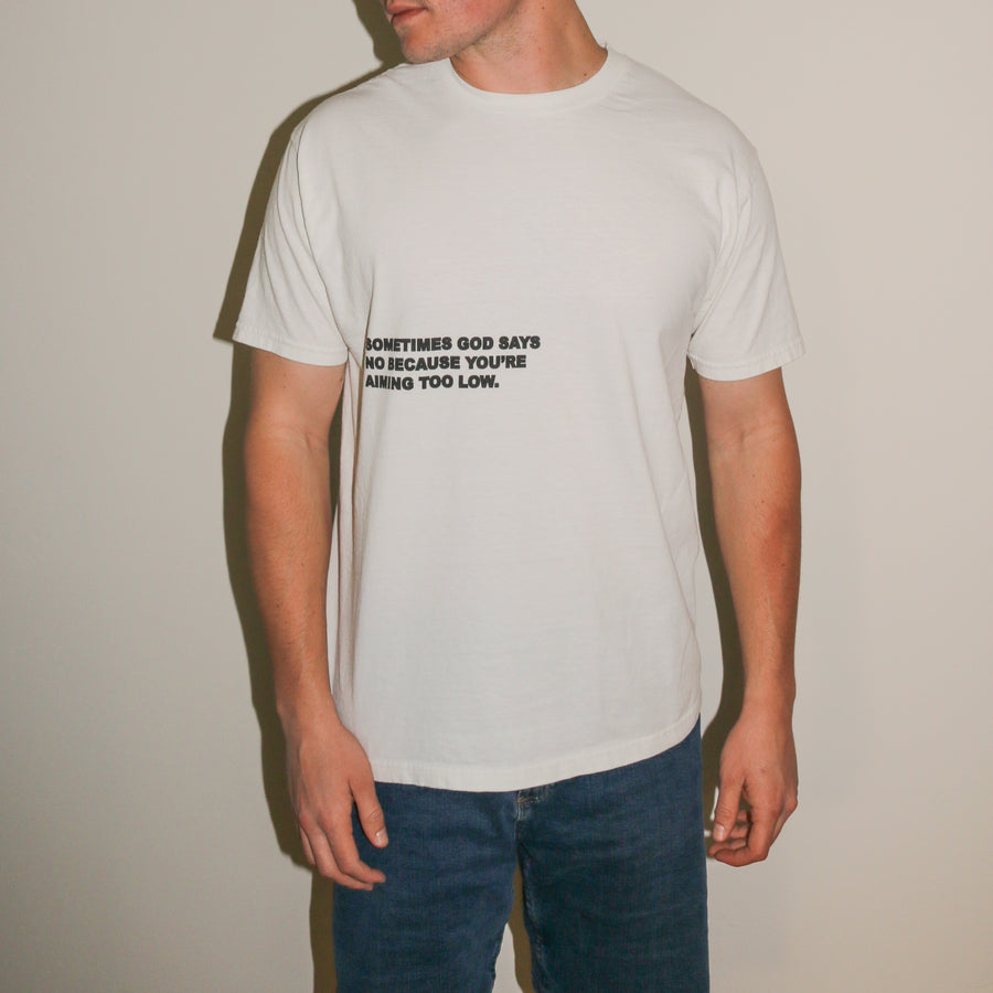 A man wearing a white crewneck t-shirt with black printed text on the chest, paired with blue jeans, standing against a plain beige wall.
