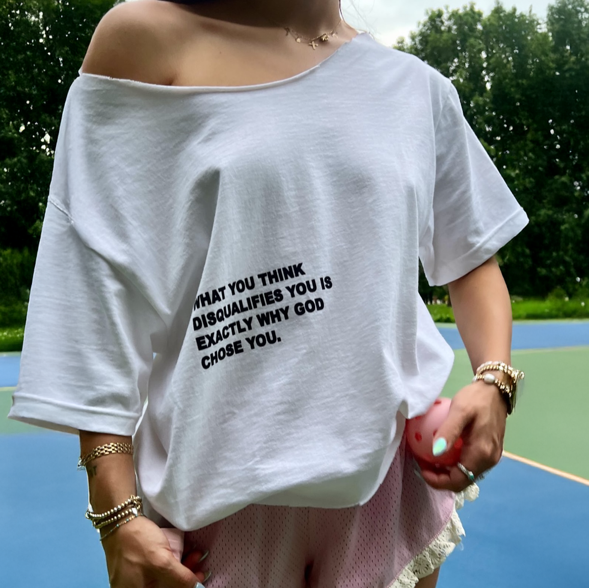 A person wearing an off-the-shoulder white graphic T-shirt with black text on the front, standing on a blue and green outdoor court with trees in the background.