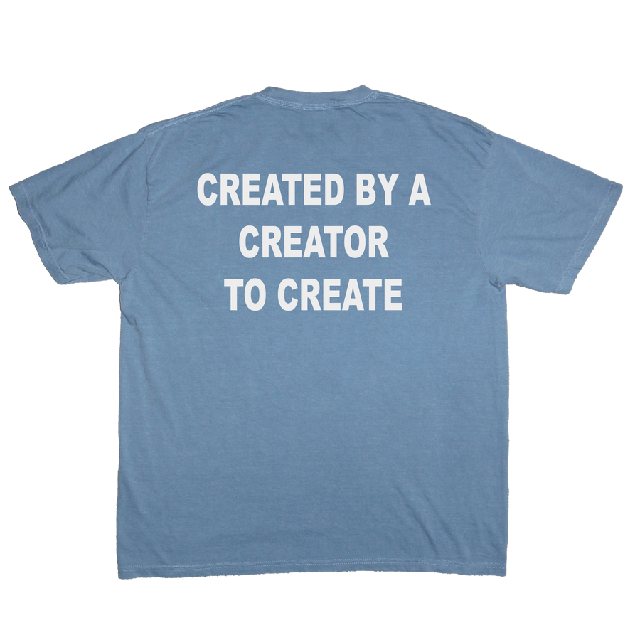 A light blue crewneck t-shirt shown from the back, featuring the phrase 'CREATED BY A CREATOR TO CREATE' printed in bold white capital letters across the upper back.